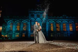 A bride and groom stand outside the illuminated Carton Towers at night, smiling as they pop a bottle of champagne on their wedding day, with sparkling bubbles and confetti flying in the air.