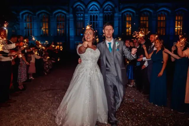 A bride and groom, smiling and holding sparklers, walk together outside at night during their wedding while guests line up on both sides. Behind them, carton towers are illuminated with blue light, creating a magical backdrop.