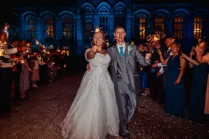 A bride and groom, smiling and holding sparklers, walk together outside at night during their wedding while guests line up on both sides. Behind them, carton towers are illuminated with blue light, creating a magical backdrop.