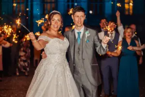 A bride and groom in wedding attire hold sparklers and smile at the camera, surrounded by guests celebrating with sparklers in the background during an enchanting outdoor wedding.