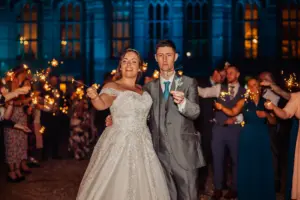 A bride and groom stand together smiling, holding sparklers at their wedding, surrounded by guests with sparklers at night, with the glowing carton towers of a large, illuminated building in the background.