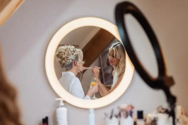 A woman applies makeup to another woman, seen through a round, illuminated mirror at Oakwood at Ryther. The bride-to-be, with styled blonde hair and a drink in hand, prepares for her Yorkshire wedding amid cosmetics and tools on the counter.