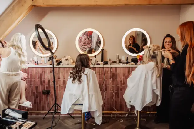 Two women in salon chairs have their hair styled by two hairstylists in front of three round lit mirrors on a pink-tiled counter—perfect Yorkshire wedding prep at Oakwood at Ryther, with beauty tools and products scattered around.