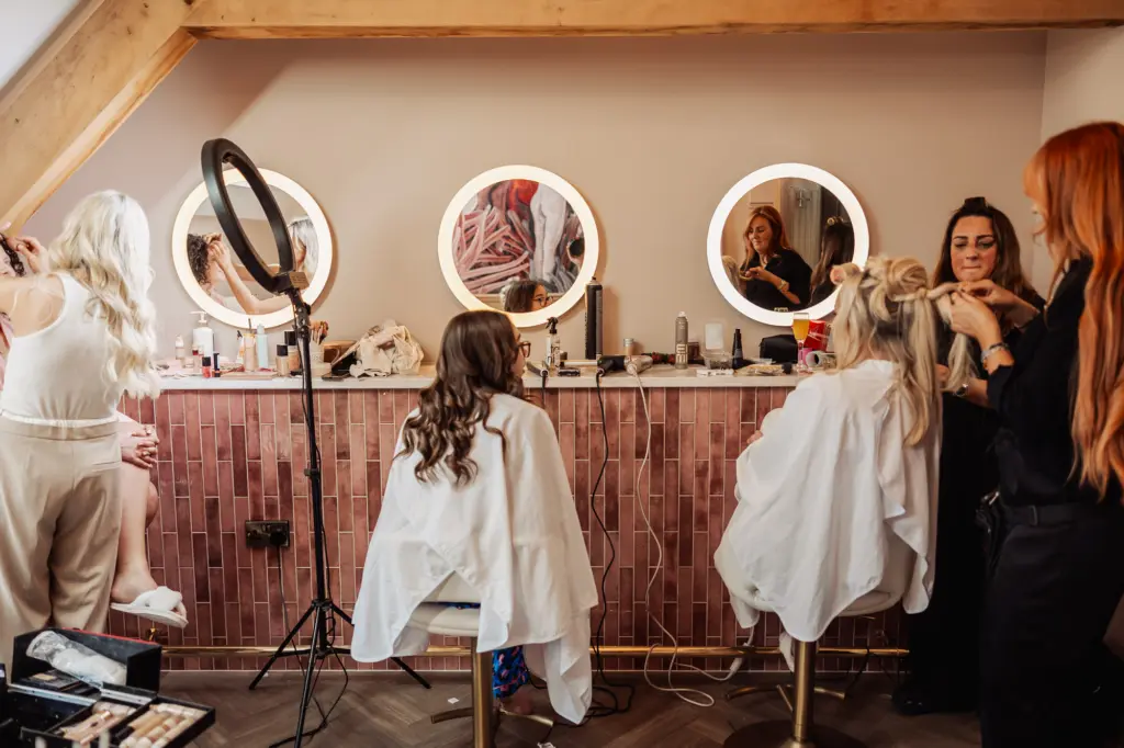 Two women in salon chairs have their hair styled by two hairstylists in front of three round lit mirrors on a pink-tiled counter—perfect Yorkshire wedding prep at Oakwood at Ryther, with beauty tools and products scattered around.
