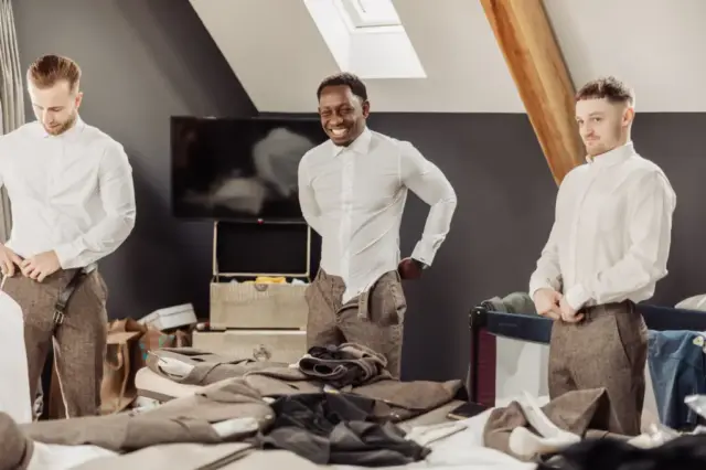 Three men in white shirts and dress pants are getting ready for a Yorkshire wedding in a modern, loft-style bedroom at Oakwood at Ryther, smiling as they adjust their clothes amid the clutter of pre-wedding excitement.