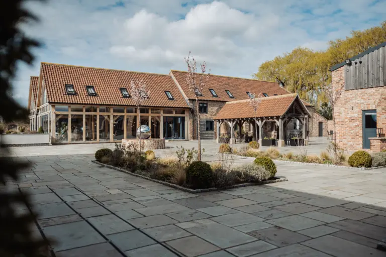 A modern barn-style building with large glass windows and a red-tiled roof sits beside a paved courtyard at Oakwood at Ryther, the perfect Yorkshire wedding venue, landscaped with small bushes and trees under a partly cloudy sky.