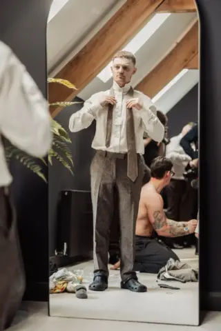 A man stands in front of a mirror adjusting his tie for a Yorkshire wedding while wearing dress pants, a white shirt, and black Crocs. Behind him at Oakwood at Ryther, others are getting ready in a room with clothes scattered on the floor.