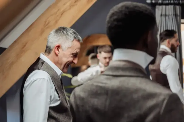 A group of men in dress shirts and vests prepare together indoors at Oakwood at Ryther, with one older man smiling in the foreground and others adjusting their outfits—a joyful moment before a Yorkshire wedding.