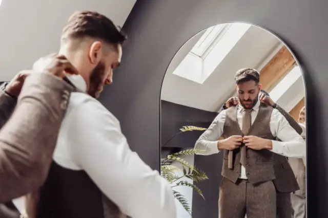 A man in a suit adjusts his vest while someone helps him from behind. He stands before a large mirror at Oakwood at Ryther, with his reflection visible in the modern, well-lit room—a perfect scene for a Yorkshire wedding.