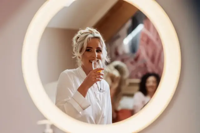 A woman in a white robe, seen through a round, illuminated mirror at Oakwood at Ryther, smiles while holding a glass of champagne. Her hair is styled up, suggesting she is preparing for her Yorkshire wedding.