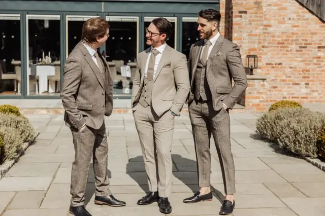 Three men in brown suits stand and chat on a paved patio at Oakwood at Ryther, with a brick building and large windows behind them. Dressed formally, they appear to be enjoying a moment together at this stylish Yorkshire wedding.