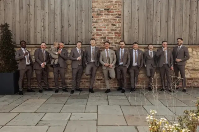 A group of ten men in matching brown suits and ties stand and lean against a stone wall outdoors at Oakwood at Ryther, smiling and posing together during a joyful Yorkshire wedding, some holding drinks and making playful gestures.