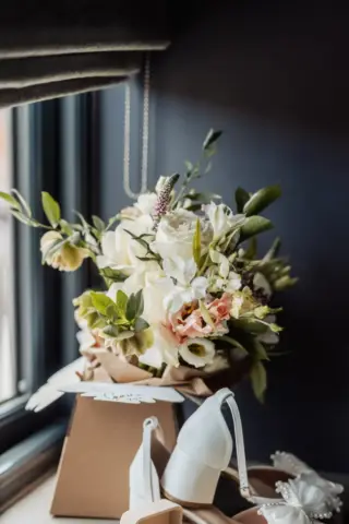 A bouquet of white and pale pink flowers with greenery sits on a windowsill at Oakwood at Ryther, alongside white bridal shoes and a brown gift box. Soft natural light completes this charming Yorkshire wedding scene.