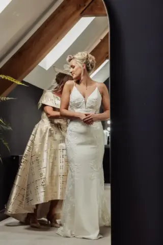 A bride in a white lace wedding dress stands in front of a mirror as another woman in a gold dress helps adjust her gown in a modern, softly lit room with exposed wooden beams at Oakwood at Ryther, the perfect Yorkshire wedding setting.