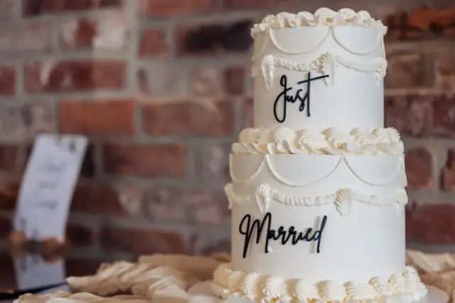 A two-tier white wedding cake with decorative icing and the words Just Married on each tier, set against a brick wall—perfect for your Oakwood at Ryther or Yorkshire wedding celebration.