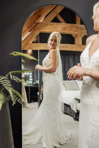 A bride in a lace wedding dress stands before a mirror at Oakwood at Ryther. Her reflection shines indoors, with wooden beams, a cozy bedroom setting, and lush greenery creating the perfect Yorkshire wedding backdrop.
