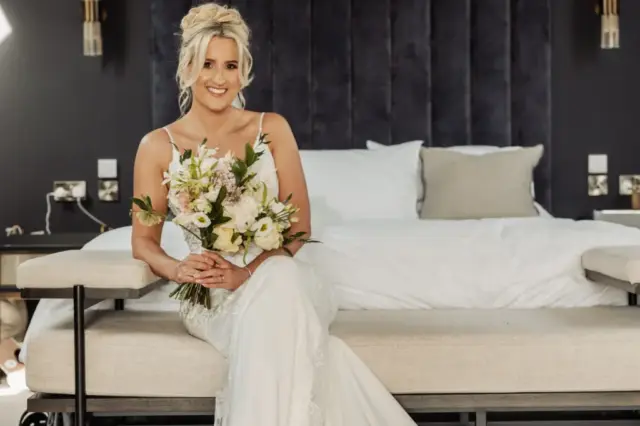 A bride in a white dress sits on a beige bench at the foot of a bed, holding a bouquet and smiling. The chic setting, with dark walls and modern lights, captures the elegance of an Oakwood at Ryther Yorkshire wedding.