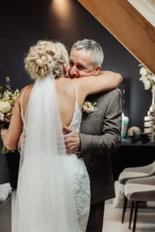 A bride in a white dress and veil hugs an older man indoors at Oakwood at Ryther. Both appear emotional and happy, surrounded by elegant modern decor. The bride holds a bouquet of flowers at this Yorkshire wedding.