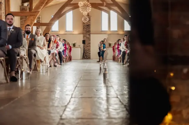 Wedding guests sit in rows on either side of a decorated aisle at Oakwood at Ryther, a rustic Yorkshire wedding venue with wooden beams and chandeliers. The foreground shows part of a wall, partially obscuring the view.