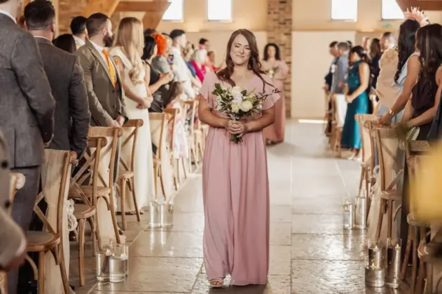 A bridesmaid in a pink dress walks down the aisle holding a bouquet, smiling, with seated guests on both sides at an Oakwood at Ryther wedding in a warmly lit indoor venue.