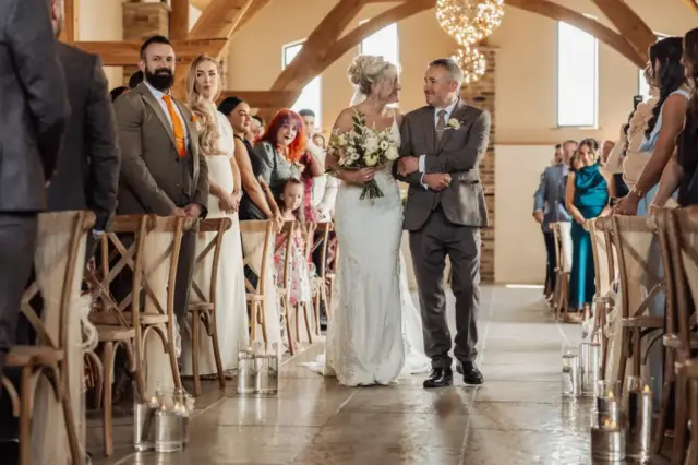 A bride in a white dress walks down the aisle with her father as smiling guests look on inside Oakwood at Ryther, creating a memorable Yorkshire wedding in a beautifully rustic, decorated venue.