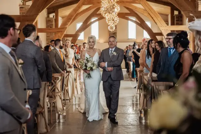 A bride in a white dress walks down the aisle with an older man in a suit, surrounded by guests in a rustic, wooden-beamed Yorkshire wedding venue, Oakwood at Ryther, beautifully decorated with candles and flowers.