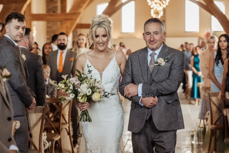 A smiling bride in a white dress holds a bouquet as she walks down the aisle arm-in-arm with an older man in a grey suit, surrounded by guests in the bright, rustic Oakwood at Ryther Yorkshire wedding venue.