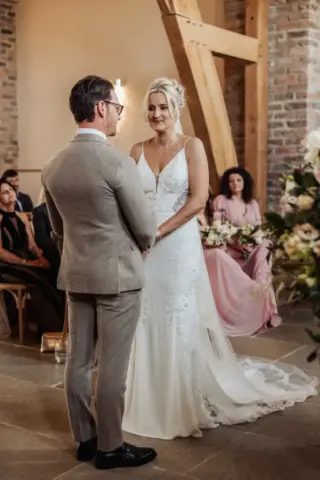 A bride and groom stand facing each other, holding hands during their wedding ceremony at Oakwood at Ryther, a rustic Yorkshire wedding venue with brick walls and wooden beams. Guests, including bridesmaids in pink dresses, are seated and watching.