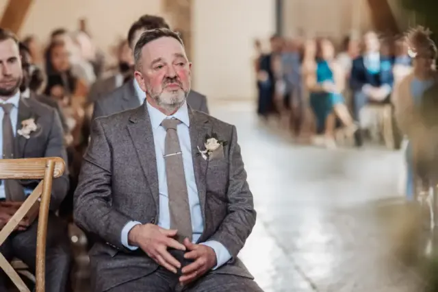 A middle-aged man in a grey suit with a boutonnière sits on a wooden chair at an Oakwood at Ryther wedding ceremony, looking emotional and teary-eyed, while guests are seated in the background.