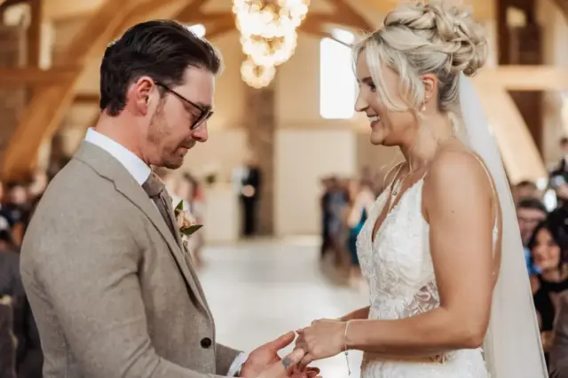 A bride and groom stand facing each other during their wedding ceremony at Oakwood at Ryther, smiling as the bride places a ring on the groom’s finger in a warmly lit venue with wooden beams and chandeliers.