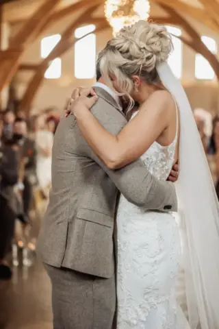 A bride and groom embrace during their wedding ceremony at Oakwood at Ryther, a sunlit Yorkshire wedding venue with arched wooden beams. The bride wears a white lace dress and veil, while the groom is in a beige suit.