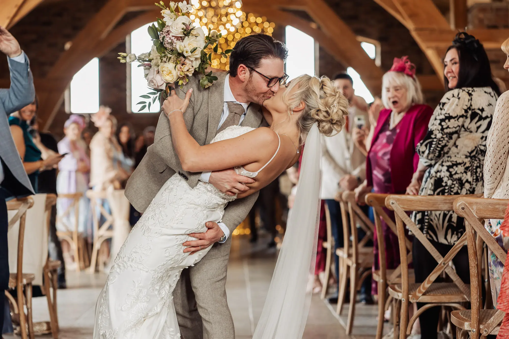 A groom in a beige suit passionately dips and kisses his bride, dressed in white gown and veil, as wedding guests cheer and capture the moment on their phones at their Yorkshire wedding in the warmly lit Oakwood at Ryther venue.