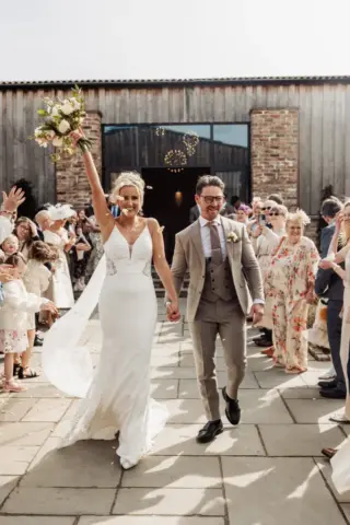 A joyful bride in a white dress raises her bouquet while holding hands with the groom in a beige suit as they walk outside at their Oakwood at Ryther Yorkshire wedding, surrounded by cheering guests at a rustic outdoor celebration.