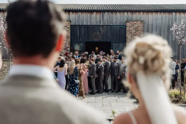 A bride and groom, seen from behind, look at a large group of wedding guests gathered outside Oakwood at Ryther on a sunny day, capturing the charm of a Yorkshire wedding.