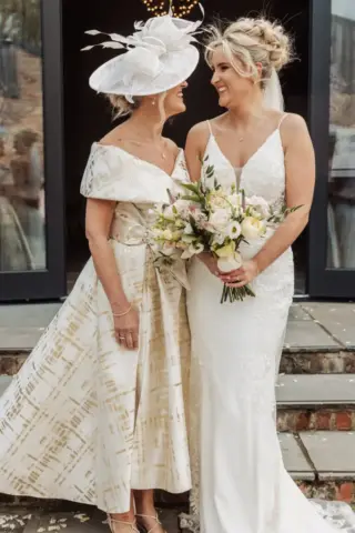 A bride in a white lace gown holding a bouquet stands smiling next to a woman in an off-shoulder dress with a large white hat. They share a joyful moment outside Oakwood at Ryther, capturing the spirit of a beautiful Yorkshire wedding.