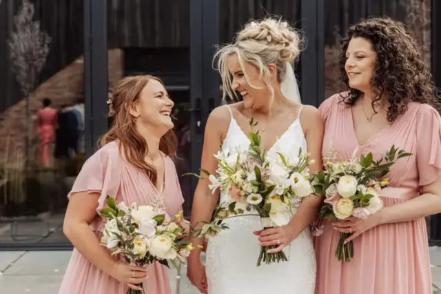 Three women in pink and white dresses, holding bouquets, smile and laugh together in front of a glass building at Oakwood at Ryther—a joyful moment from a Yorkshire wedding, with the bride flanked by her two bridesmaids.
