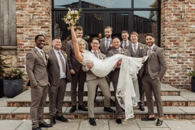 A bride in a white dress is held up by a groom, surrounded by eight groomsmen in brown suits, all smiling and posing at an Oakwood at Ryther Yorkshire wedding. The bride holds her bouquet triumphantly in the air.