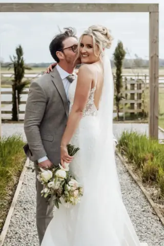 A groom in a grey suit kisses a smiling bride in a lace wedding dress holding a bouquet, standing together outdoors on a gravel path with greenery and wooden fencing at Oakwood at Ryther, the perfect Yorkshire wedding setting.