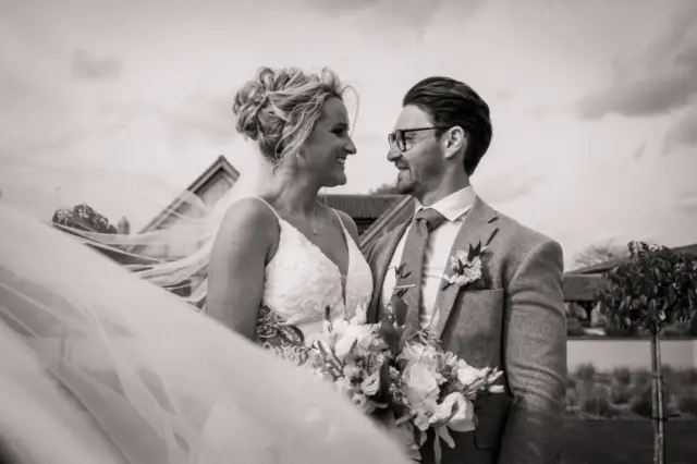 A bride and groom stand close together outdoors at Oakwood at Ryther, smiling and gazing into each other’s eyes. The bride holds a bouquet and flowing veil, while the groom wears glasses and a suit with a boutonniere—perfect Yorkshire wedding moments.
