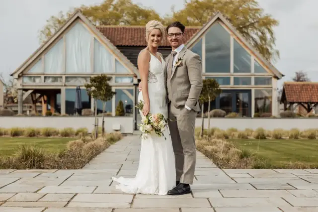 A bride in a white dress and a groom in a light grey suit smile together outside Oakwood at Ryther, holding a bouquet on a stone pathway lined with grass and shrubs, capturing the essence of a beautiful Yorkshire wedding.
