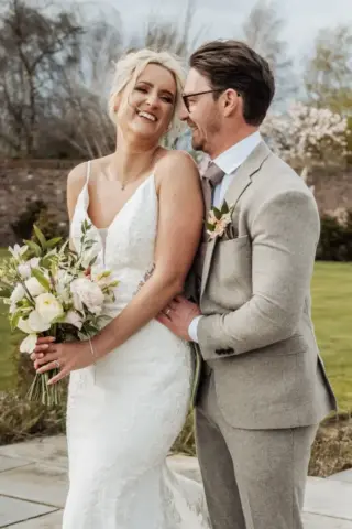A bride in a white lace dress holding a bouquet smiles joyfully while her groom in a light gray suit and glasses stands beside her, holding her waist. They stand outdoors at Oakwood at Ryther, surrounded by lush greenery—a perfect Yorkshire wedding.