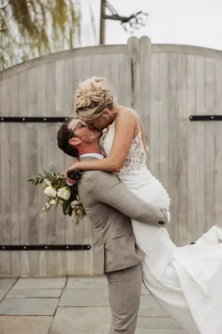 A groom in a light gray suit lifts and kisses the bride, who is holding a bouquet and wearing a lace wedding dress, in front of rustic wooden gates at their Oakwood at Ryther Yorkshire wedding.