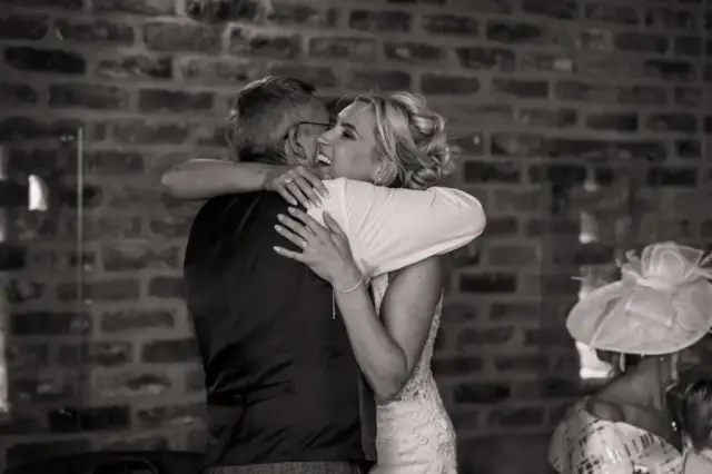 A joyful bride in a wedding dress hugs an older man in a vest as they smile at a Yorkshire wedding. A woman in a hat sits nearby, framed by a brick wall. The black and white image captures Oakwood at Ryther’s timeless charm.