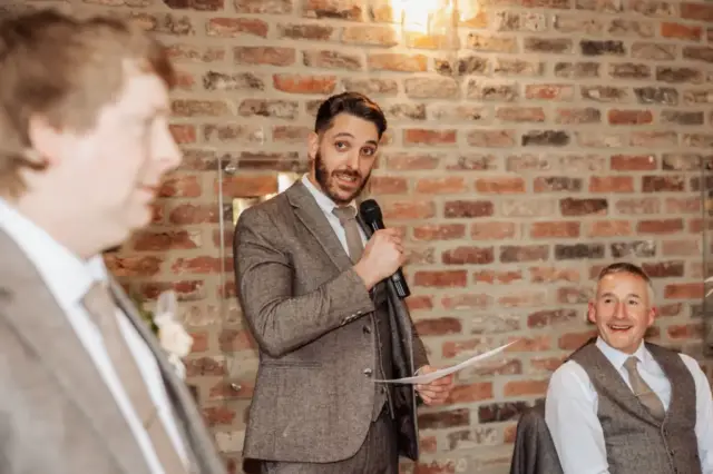 A man in a gray suit speaks into a microphone while holding papers, standing between two seated men in suits, all against a brick wall background at an Oakwood at Ryther Yorkshire wedding. The atmosphere appears formal and cheerful.
