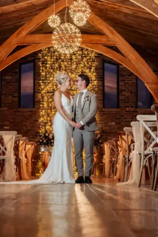 A bride and groom stand holding hands in a rustic wedding venue at Oakwood at Ryther, with wooden beams, fairy lights, and hanging globe lights. Rows of decorated chairs line the aisle as the couple smiles, capturing a perfect Yorkshire wedding moment.