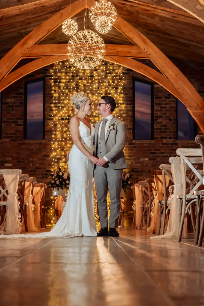A bride and groom stand holding hands in a rustic wedding venue at Oakwood at Ryther, with wooden beams, fairy lights, and hanging globe lights. Rows of decorated chairs line the aisle as the couple smiles, capturing a perfect Yorkshire wedding moment.