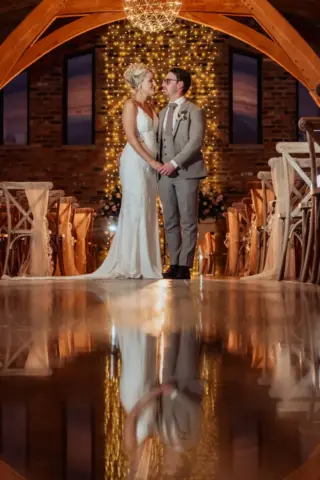 A bride and groom stand holding hands in a warmly lit aisle at Oakwood at Ryther, decorated with fairy lights and wooden arches, their reflection visible on the polished floor beneath them—capturing the magic of a Yorkshire wedding.