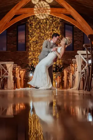 A groom dips his bride for a kiss on a polished floor inside Oakwood at Ryther, a rustic Yorkshire wedding venue with string lights and wooden beams overhead, creating a romantic, glowing atmosphere.