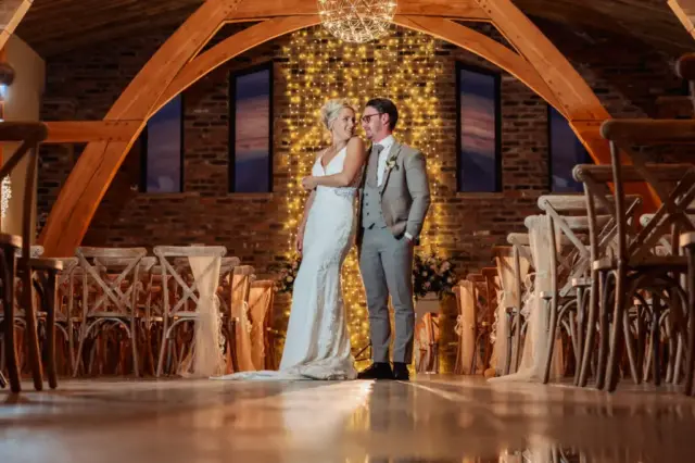 A bride and groom stand closely together in an elegant wooden hall at Oakwood at Ryther, decorated with fairy lights and empty chairs, sharing a joyful, intimate moment on their Yorkshire wedding day.