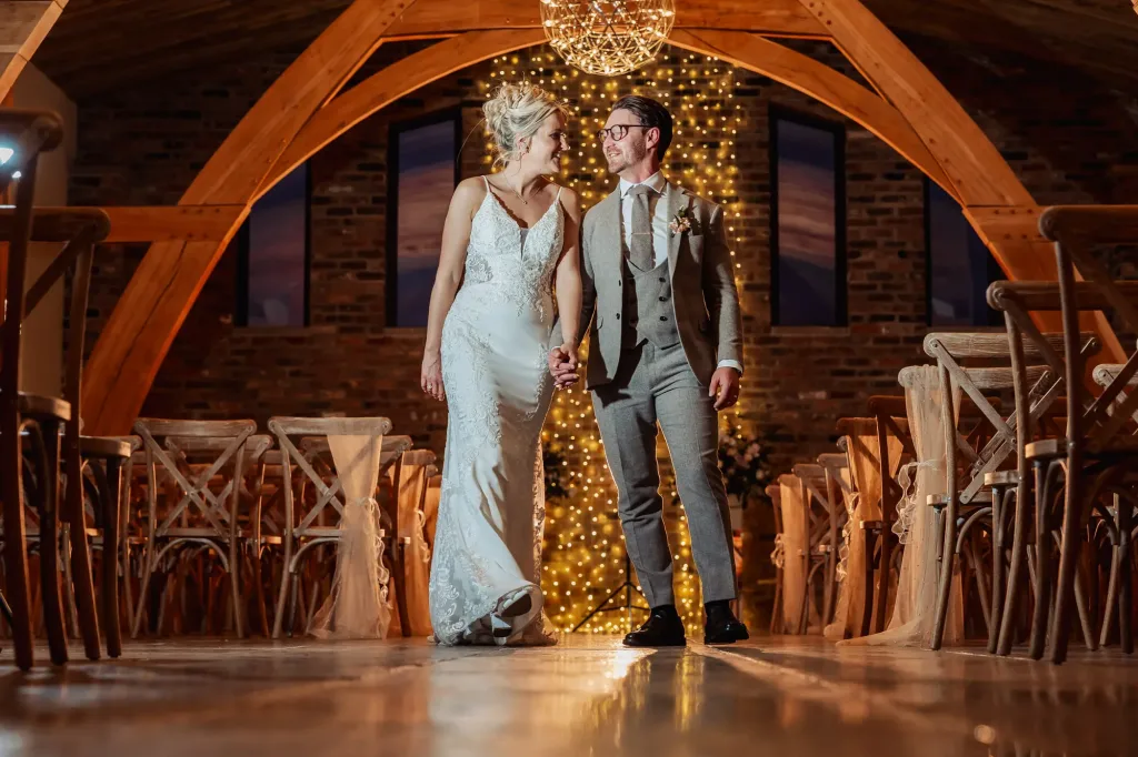 A bride and groom holding hands and smiling at each other as they walk down the aisle in Oakwood at Ryther, a beautifully decorated Yorkshire wedding venue with warm lighting and string lights glowing behind them.